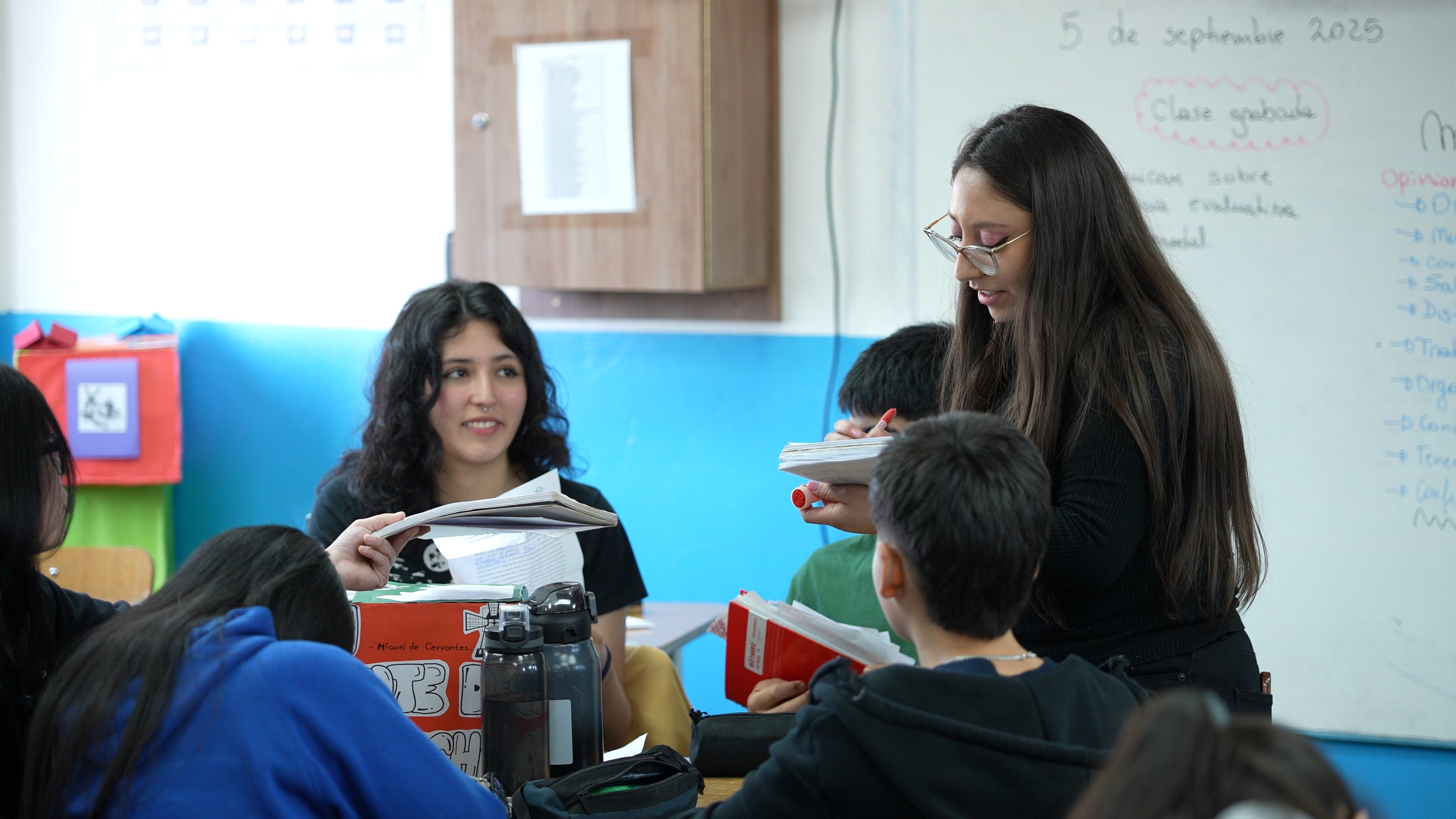 A teacher and student lean over a desk to assist a group of children working together in a classroom.