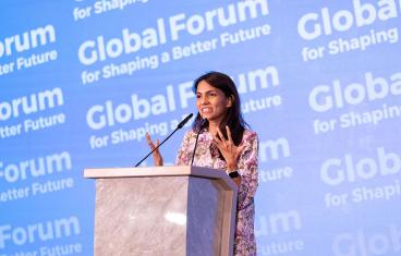 A young woman with long brown hair and tan skin stands at a podium in front of a background that says Global Forum for Shaping a Better Future