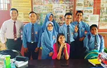 An Asian woman in a pink shirt smiles while seated at table surrounded by teenage students in blue uniforms