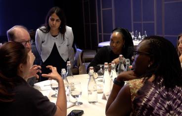 A woman with long brown hair wearing a white blazer leans over a table of diverse adults who appear to be in discussion