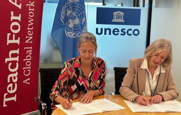 Two white women with greying blonde hair sit side by side signing papers in front of them, a red Teach For All banner on the right, and a UNESCO logo above one's head