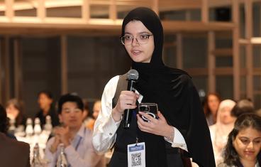 A young woman wearing a black head scarf and glasses speaks into a microphone 