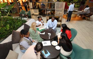 Diverse professionals gather around small tables in a modern lounge for a collaborative networking session featuring snacks, drinks, and casual discussion.