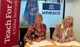 Two white women with greying blonde hair sit side by side signing papers in front of them, a red Teach For All banner on the right, and a UNESCO logo above one's head
