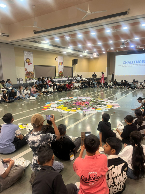 A large group of students sits in a wide circle on an auditorium floor around a colorful, circular arrangement of papers and artwork.