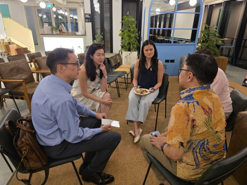 Four people sit in a circle on chairs in a modern office, engaged in an earnest conversation while one woman holds a plate of food.