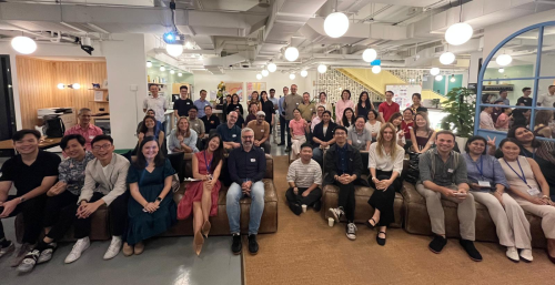 A large, diverse group of professionals smiles for a group photo in a brightly lit, modern industrial-style office space with exposed ceilings and warm lighting.