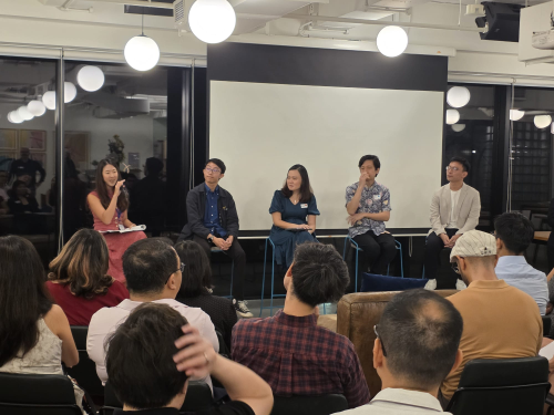 Five speakers sit on chairs facing an audience during a panel discussion in a modern office space with large windows and a projector screen.