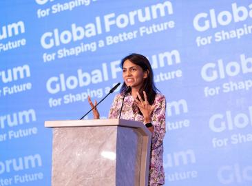 A young woman with long brown hair and tan skin stands at a podium in front of a background that says Global Forum for Shaping a Better Future