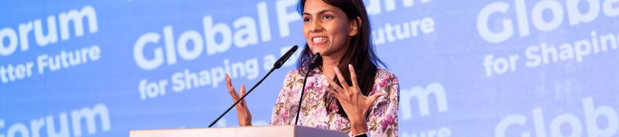 A young woman with long brown hair and tan skin stands at a podium in front of a background that says Global Forum for Shaping a Better Future