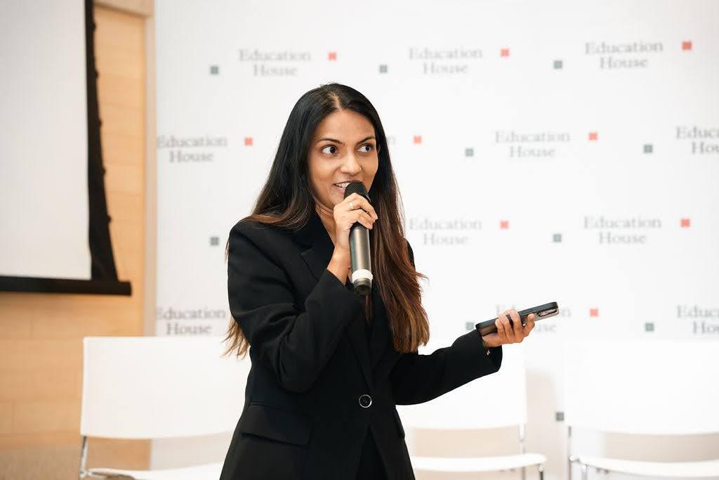 Cheryl Fernando holds a microphone and gestures with a remote while speaking in front of a backdrop featuring "Education House" logos during a presentation.