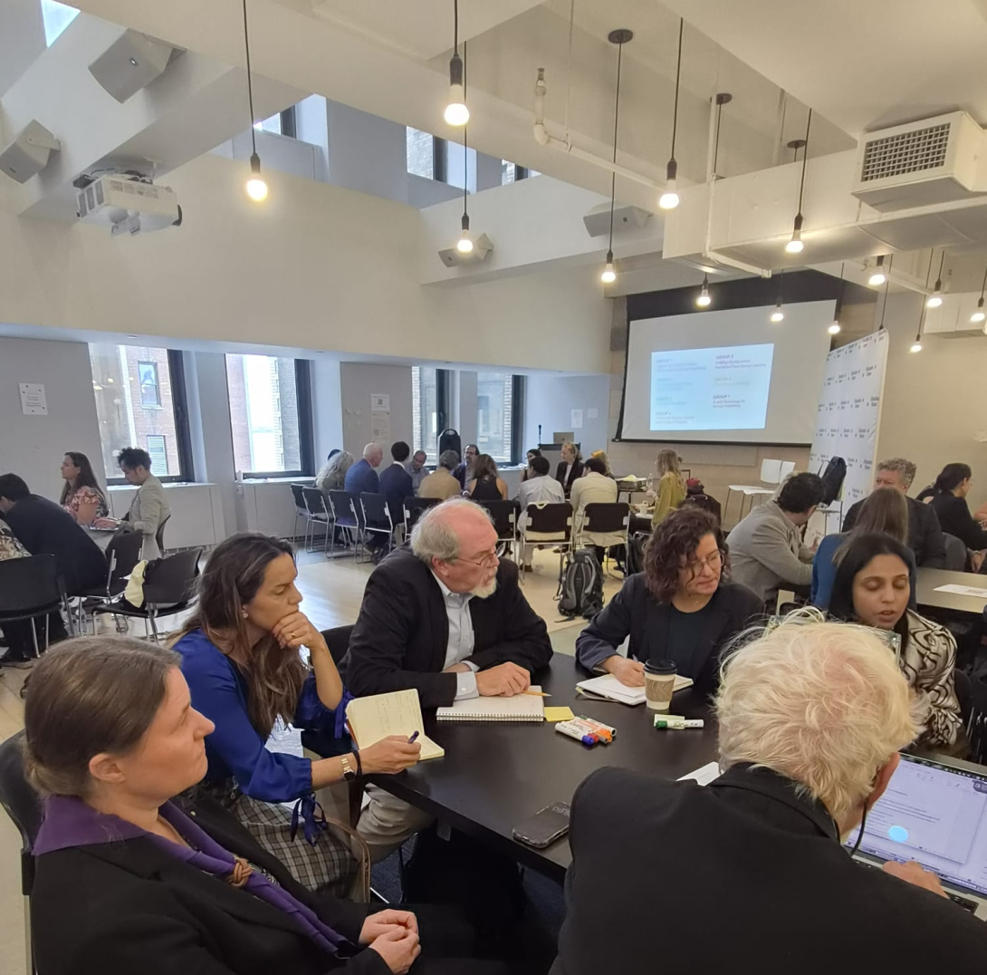 People seated around a dark table converse during a workshop, while other attendees gather at small tables in a bright, modern room.